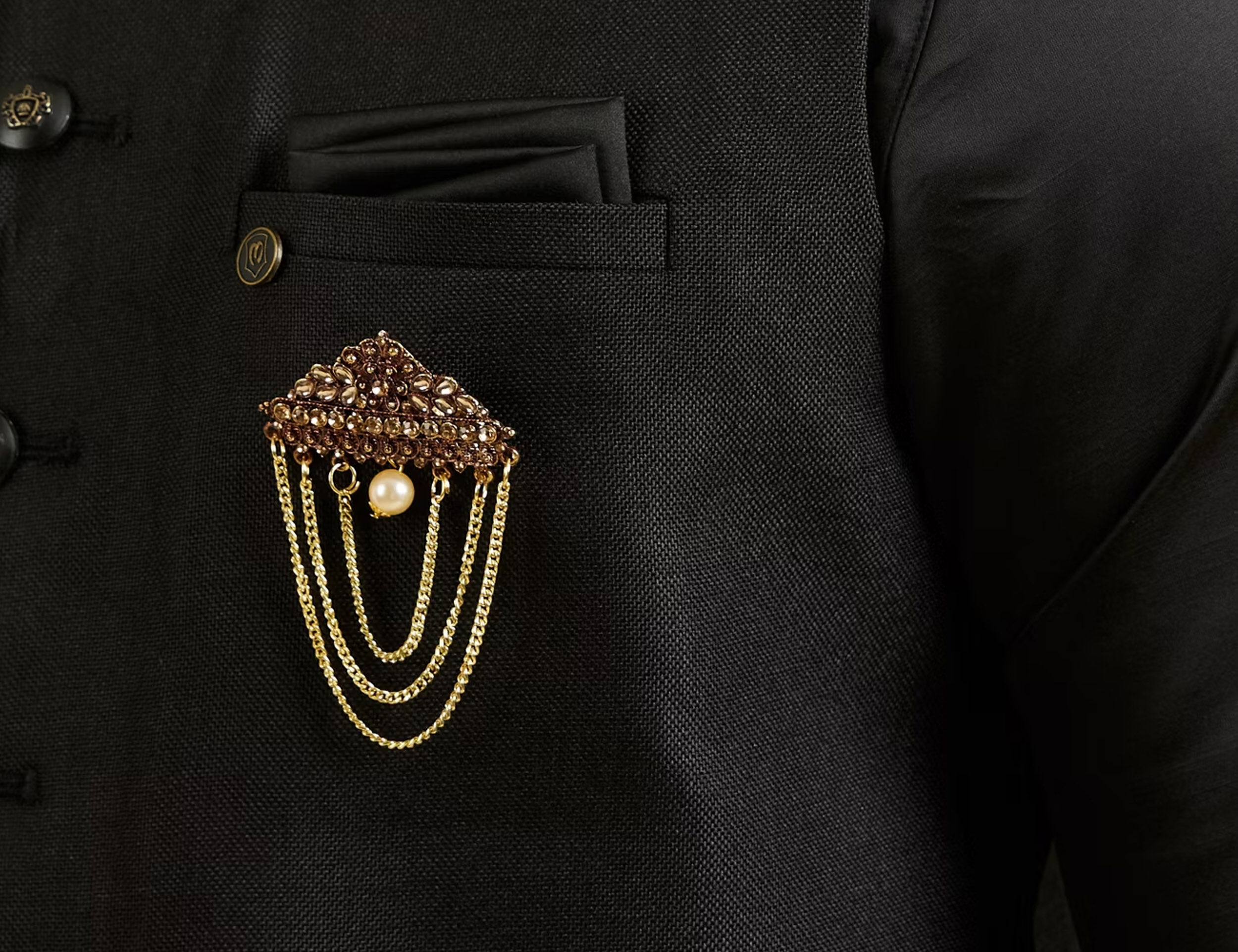 Gold chained brooch with a central pearl and jeweled triangle design pinned to the lapel of a black suit.